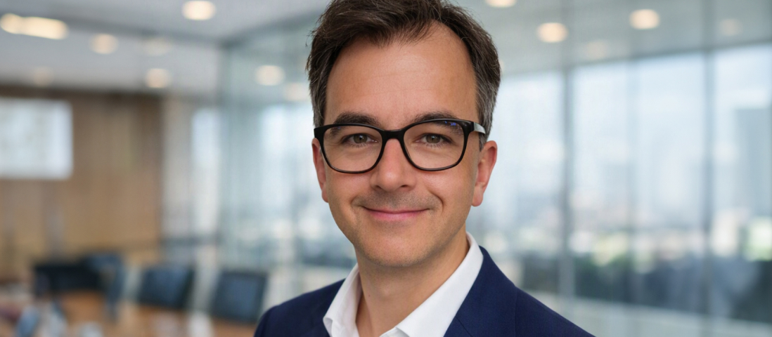 Professional headshot of a person wearing glasses, a white shirt, and a blue blazer, standing in a modern office meeting room.